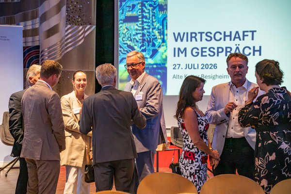 Eine Gruppe von Fachleuten steht in einem Raum und diskutiert lebhaft. Im Hintergrund ist ein Bildschirm mit der Aufschrift „Wirtschaft im Gespräch“ und dem Datum 27. Juli 2026 zu sehen. Die Atmosphäre ist informell und fördert den Austausch von Ideen.