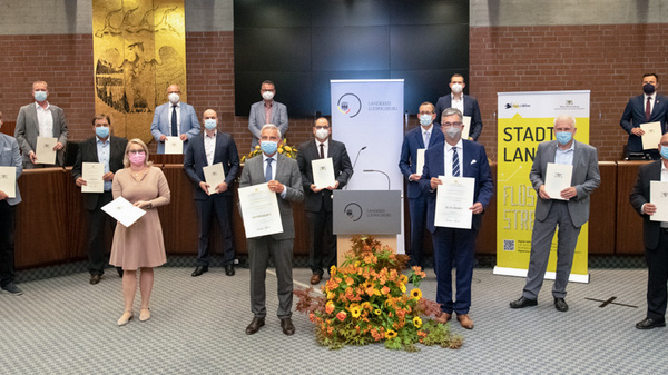 Un groupe de personnes posant ensemble dans une salle de réunion, tenant des certificats. Tous portent des masques. L'atmosphère est formelle et engageante, avec des éléments de décoration florale en avant-plan et des bannières affichant des messages sur le mur.