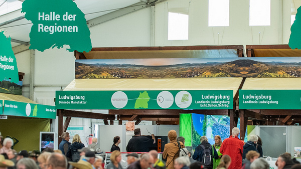 Un intérieur de hall d'exposition avec de nombreuses personnes. Des stands, portant des panneaux verts, sont visibles à l'arrière-plan, avec des inscriptions en allemand mentionnant "Halle der Regionen". La foule s'active autour des différents kiosques. 

(Text KI generiert)