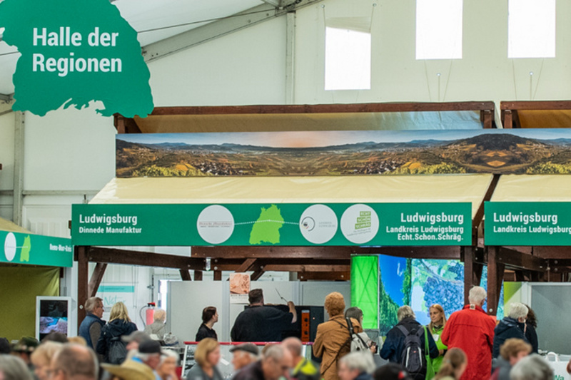 Ein überfüllter Veranstaltungsraum mit Ständen und Anzeigetafeln, die die "Halle der Regionen" markieren. Die Menschen stehen zusammen, während einige an den Ständen einkaufen oder informieren. Helle Beleuchtung und ein großes Zeltdach sind sichtbar. (Text KI generiert)