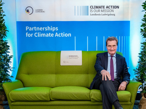 Ein Mann sitzt auf einer grünen Couch vor einem blauen Hintergrund mit dem Text "Climate Action is our mission" und "Partnerships for Climate Action". Die Umgebung ist mit Pflanzen dekoriert, was eine einladende Atmosphäre schafft.