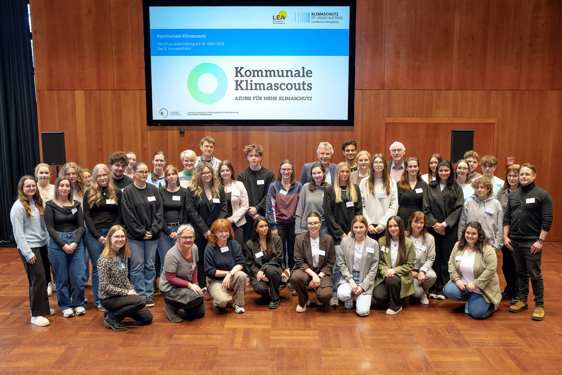 Eine große Gruppe von Menschen steht in einem Raum und posiert für ein Foto. Im Hintergrund ist ein Bildschirm mit dem Text "Kommunale Klimascouts - Aktive für mehr Klimaschutz" zu sehen. Die Teilnehmerinnen und Teilnehmer sind gemischt zwischen verschiedenen Altersgruppen und Geschlechtern.