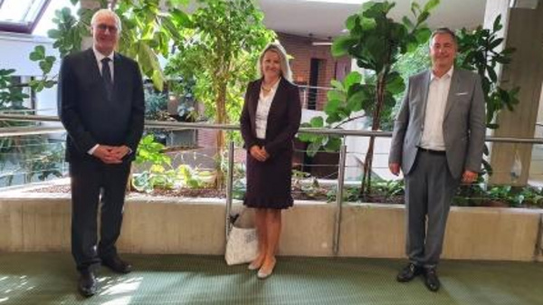Three individuals stand in a well-lit indoor space. Two men, one in a dark suit and the other in a light gray suit, are on either side of a woman in a dark dress coat. Surrounding them are various indoor plants, contributing to a green ambiance.