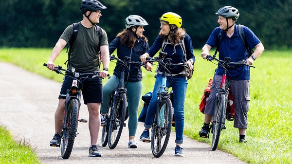 Vier Personen auf Fahrrädern stehen auf einem Weg in einer grünen Landschaft. Zwei Männer und zwei Frauen lachen und unterhalten sich. Alle tragen Helme und sportliche Kleidung. Das Bild vermittelt eine freundliche, gesellige Atmosphäre im Freien.