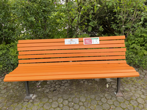 Panchina di legno arancione situata in un'area verde. Presenta due cartelli informativi attaccati sullo schienale, uno dei quali è rosso. Lo sfondo mostra fogliame verde.