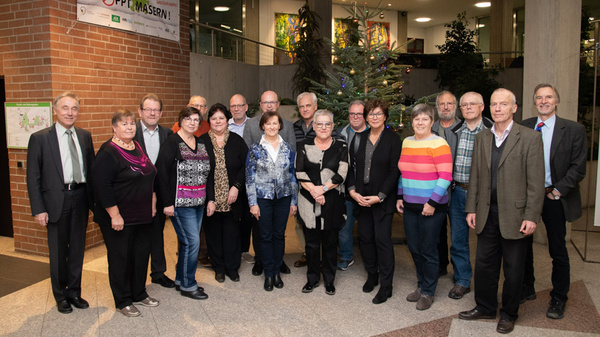 A group of 20 people stands together in a well-lit indoor space, with a Christmas tree in the background. They are dressed in various colors and styles, smiling at the camera. A banner reading "STOPP MASERN!" is visible above them.