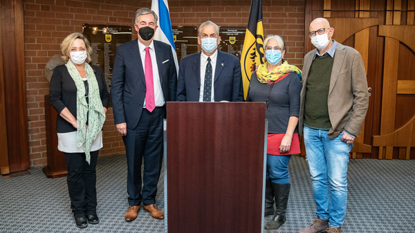 Cinq personnes se tiennent devant un pupitre. Elles portent des masques et affichent des expressions sérieuses. De drapeaux en arrière-plan suggèrent un cadre officiel. L'image dénote un événement important ou une conférence de presse.