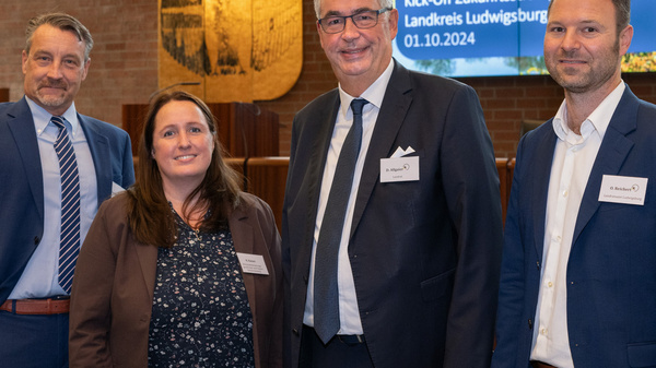 Vier Personen stehen zusammen in einem Raum, der für eine Veranstaltung vorbereitet ist. Im Hintergrund ist ein Bildschirm mit dem Text „Herzlich Willkommen“ und Informationen zu einer Kick-Off-Veranstaltung für die Zukunftsstrategie des Landkreises Ludwigsburg am 01.10.2024.