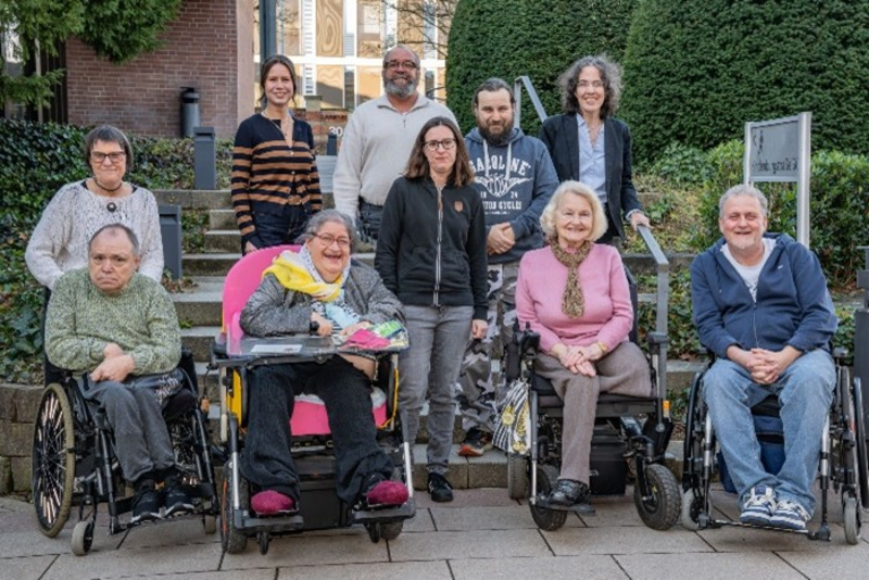 Eine Gruppe von neun Menschen, darunter vier Personen im Rollstuhl, posiert draußen vor einer Gebäudefassade. Im Hintergrund sind einige Bäume und ein Gehweg sichtbar. Alle Anwesenden lächeln und scheinen eine freundliche Atmosphäre zu schaffen.