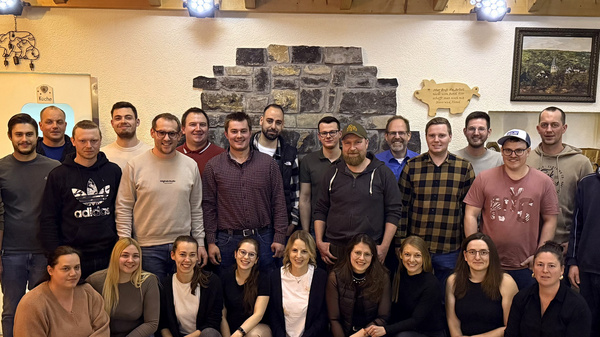 Eine Gruppe von etwa 30 Personen steht in einem Raum mit Holzbalkendecke. Sie posieren gemeinsam für das Foto, lächeln und wirken freundlich. Im Hintergrund sind Steinwände und einige Dekorationselemente sichtbar. Die Menschen sind in verschiedenen Kleidungsstilen gekleidet.