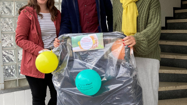 Drei Personen stehen in einem Hausflur vor einem Kinderwagen, der mit Luftballons und einigem Geschenkpapier dekoriert ist. Die Frauen tragen unterschiedliche Oberbekleidung. Der Mann in der Mitte lächelt und hält den Kinderwagen fest. Es scheint eine Geschenkübergabe oder Feierlichkeit zu sein.