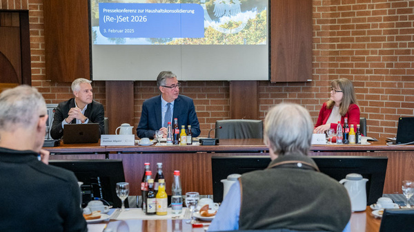 Ein Konferenzraum mit mehreren Personen, die an einem Tisch sitzen. Vorne sind drei Redner: ein Mann in Anzug, ein zweiter Mann und eine Frau mit langen Haaren. Im Hintergrund ist ein Bildschirm mit der Aufschrift "Pressekonferenz zur Haushaltskonsolidierung (Re-)Set 2026". Getränke und Snacks sind auf dem Tisch verteilt.