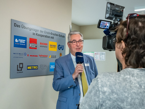 08.04.2025 - Interview zur Einweihung des Übungslagers an der Erich-Bracher-Schule Ein Mann mit Brille und Anzug steht an einem Schild, das Informationen über ein Übungslager der Erich-Bracker-Schule enthält. Er hält ein Mikrofon und spricht mit einer Interviewerin, die eine Kamera hält. Hinter ihnen ist ein heller Raum zu sehen.