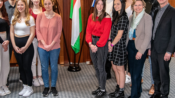 Eine Gruppe von jungen Menschen und Erwachsenen steht in einem Raum, umgeben von Flaggen. Sie lächeln und posieren gemeinsam für ein Gruppenfoto. Der Hintergrund zeigt Holzwände und ein moderner Bodenbelag.