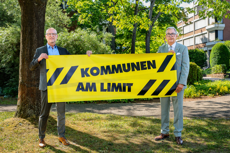 Zwei Männer stehen im Freien und halten ein großes, gelbes Banner mit der Aufschrift "KOMMUNEN AM LIMIT". Im Hintergrund sind Bäume und ein Gebäude sichtbar. Die Botschaft hebt die Herausforderungen hervor, mit denen Kommunen konfrontiert sind.
