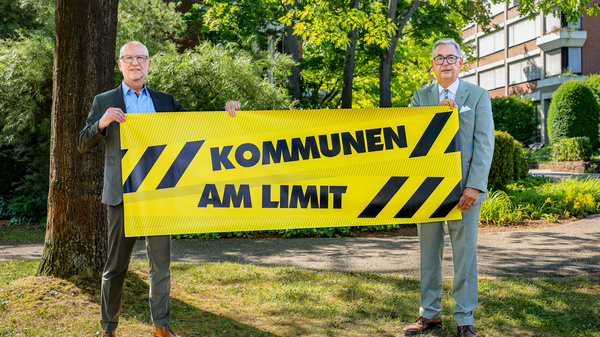 Zwei Männer stehen im Freien und halten ein großes, gelbes Banner mit der Aufschrift "KOMMUNEN AM LIMIT". Im Hintergrund sind Bäume und ein Gebäude sichtbar. Die Botschaft hebt die Herausforderungen hervor, mit denen Kommunen konfrontiert sind.