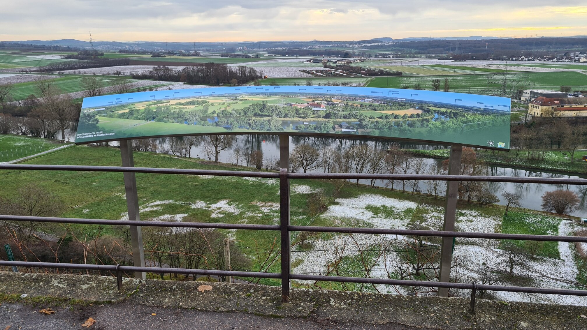 Ein Panoramablick auf eine grüne Landschaft mit Feldern, einem Fluss und mehreren Bäumen. Im Vordergrund ist eine Informationstafel mit einer Karte oder Darstellung der Umgebung. Der Himmel ist bewölkt, und es herrscht eine ruhige, natürliche Atmosphäre.