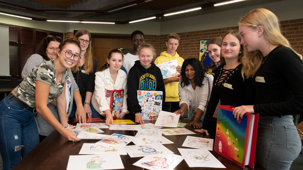 A group of teenagers stands around a table filled with colorful drawings. They smile and pose together, showcasing their artwork. Some hold additional sheets and portfolios. The setting appears to be an indoor space, possibly a classroom or community center.