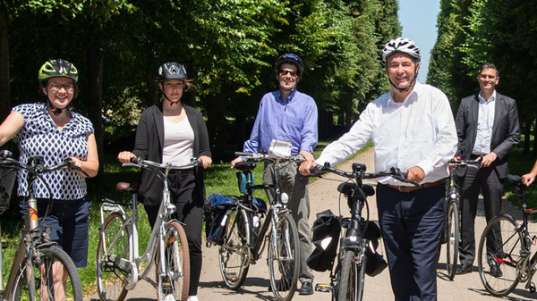A group of seven individuals, wearing helmets and standing next to their bicycles, poses together on a sunny pathway lined with trees. They appear to be participating in a cycling event or promotion, showcasing camaraderie and support for cycling activities.