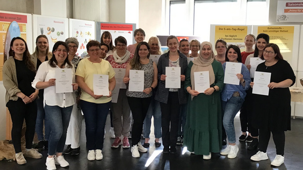 Eine Gruppe von etwa 20 Menschen steht in einem Raum und hält Urkunden in der Hand. Sie lächeln und stehen vor Informationsplakaten an der Wand. Die Atmosphäre wirkt feierlich und erfreulich.