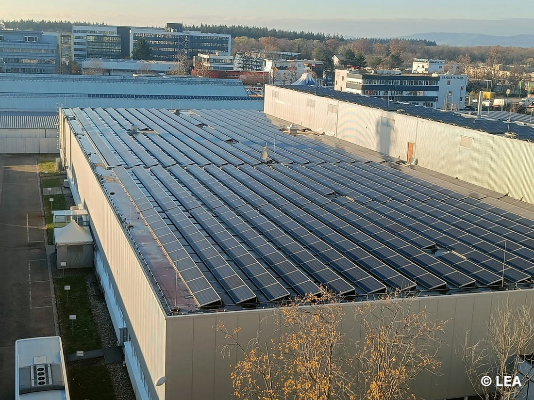 Dächer von Gebäuden mit zahlreichen Solarpanelen, die in der Sonne glänzen. Im Hintergrund sind mehrere Industriegebäude und Bäume sichtbar. (Text KI generiert)