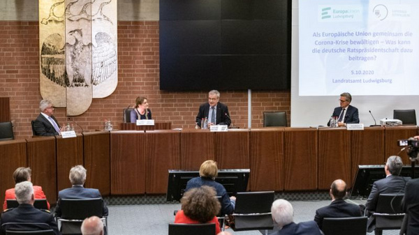 A panel discussion taking place in a conference room, featuring four speakers seated at a wooden table. A screen behind them displays text related to the European Union's response to the COVID-19 crisis, indicating the date as October 5, 2020. Audience members are seated in the background.