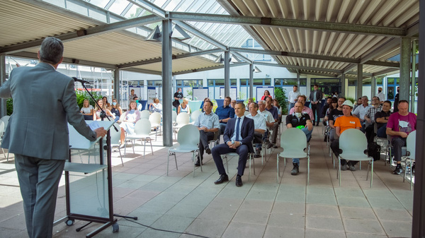 A speaker in a gray suit stands at a podium addressing an audience in a spacious, sunlit venue. The audience, seated in rows, includes individuals in various attire, some taking notes. The setting features glass panels and a modern design, enhancing the atmosphere of engagement and discussion.