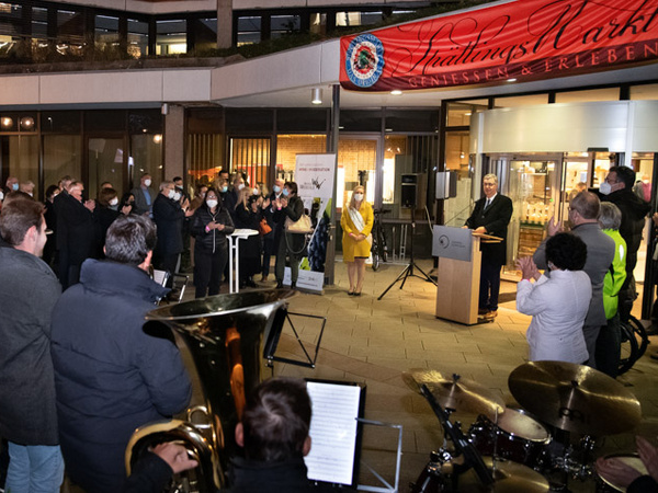 28.10.2021 - Eröffnung des Spätlingsmarkts Eine Gruppe von Menschen applaudiert bei einer Veranstaltung im Freien, während ein Redner auf einer Bühne spricht. Im Hintergrund sind Instrumente einer Band zu sehen, und ein Banner mit der Aufschrift „Hildesheimer Markt“ hängt über der Szene. Die Atmosphäre ist feierlich.