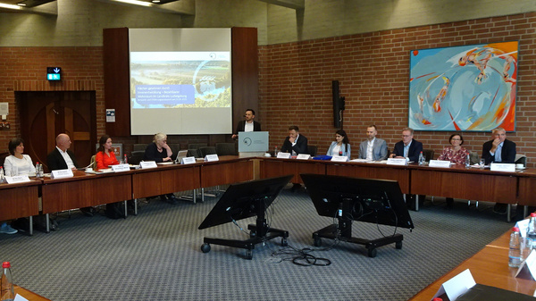 Eine Konferenz oder ein Meeting mit mehreren Teilnehmern, die um einen großen, rechteckigen Tisch sitzen. An einer Wand steht ein Präsentationsbildschirm, auf dem Informationen angezeigt werden. Ein farbenfrohes Kunstwerk hängt an der anderen Wand.
