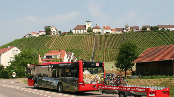 A red public bus is parked on a rural road, displaying a promotional advertisement. Attached to it is a trailer carrying bicycles and local products. In the background, rolling vineyards lead up to a small village with houses and a church on a hill.