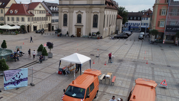 A public square featuring a church, fountain, and several vendors under tents. Two orange vehicles are parked nearby. People are scattered throughout the area, engaging in various activities amidst a backdrop of buildings and trees. The sky is partly cloudy.