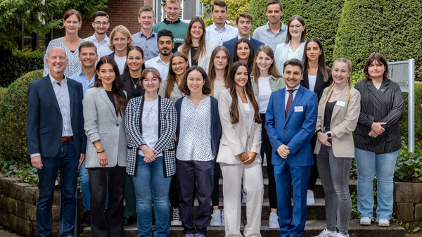 Gruppe von 24 Personen, die im Freien steht, umgeben von grünen Pflanzen. Die Personen sind in formeller und lässiger Kleidung gekleidet. Sie lächeln und posieren zusammen, wobei einige nah beieinander stehen und andere etwas Abstand halten.