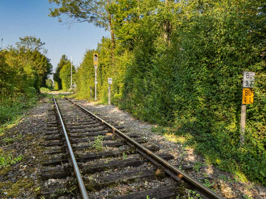 Header Bild: Ein stiller Bahngleisabschnitt führt durch dichte, grüne Bäume. Auf der rechten Seite stehen zwei Schilder, die auf Bahnübergänge hinweisen. Die Umgebung wirkt friedlich und unberührt, während die Gleise in die Ferne verlaufen. (Text KI generiert)