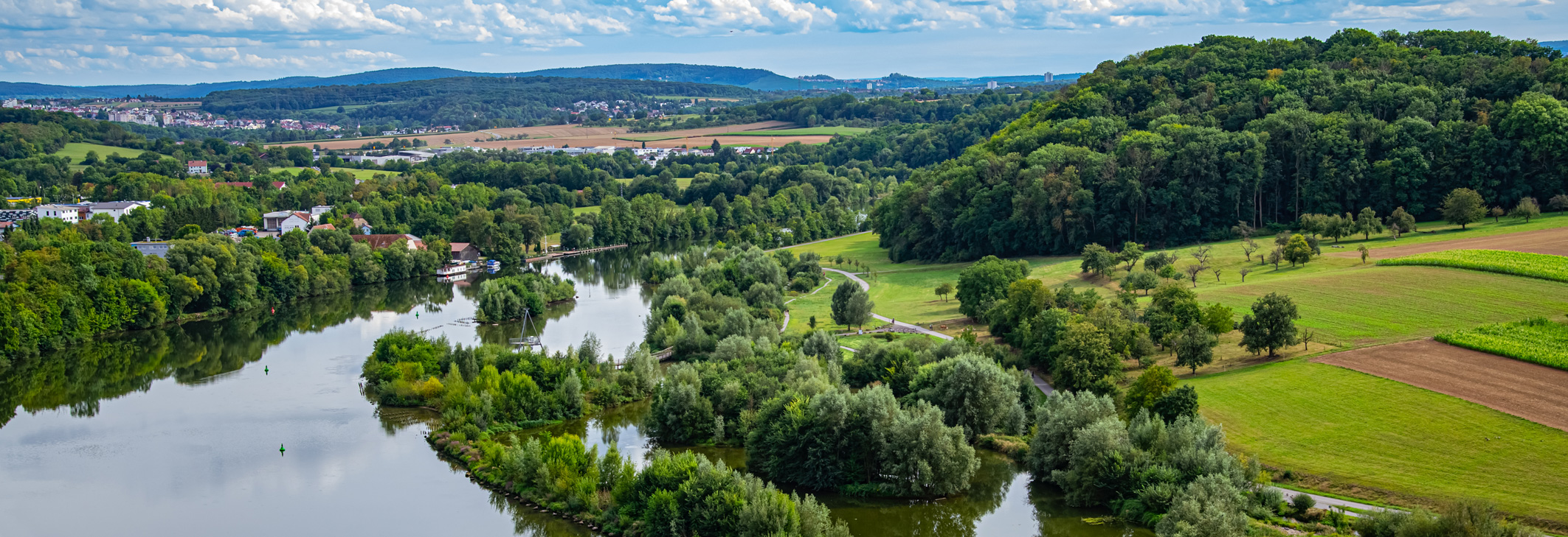 Header Bild: Eine ruhige Flusslandschaft mit grünen Ufern und Bäumen, die sich an das Wasser schmiegen. Im Hintergrund erstreckt sich sanftes Hügelland unter einem bewölkten Himmel. Kleine Ortschaften sind in der Ferne sichtbar, umgeben von Feldern und Wiesen.