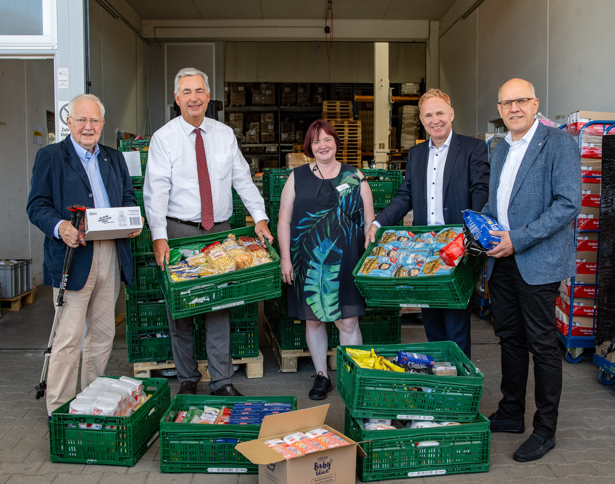 Fünf Personen stehen in einem Lagerraum und halten Korb mit Lebensmitteln. Alle lachen und wirken freundlich. Im Hintergrund sind weitere Kisten mit Lebensmitteln zu sehen. Die Szene vermittelt eine unterstützende und gemeinschaftliche Atmosphäre.
