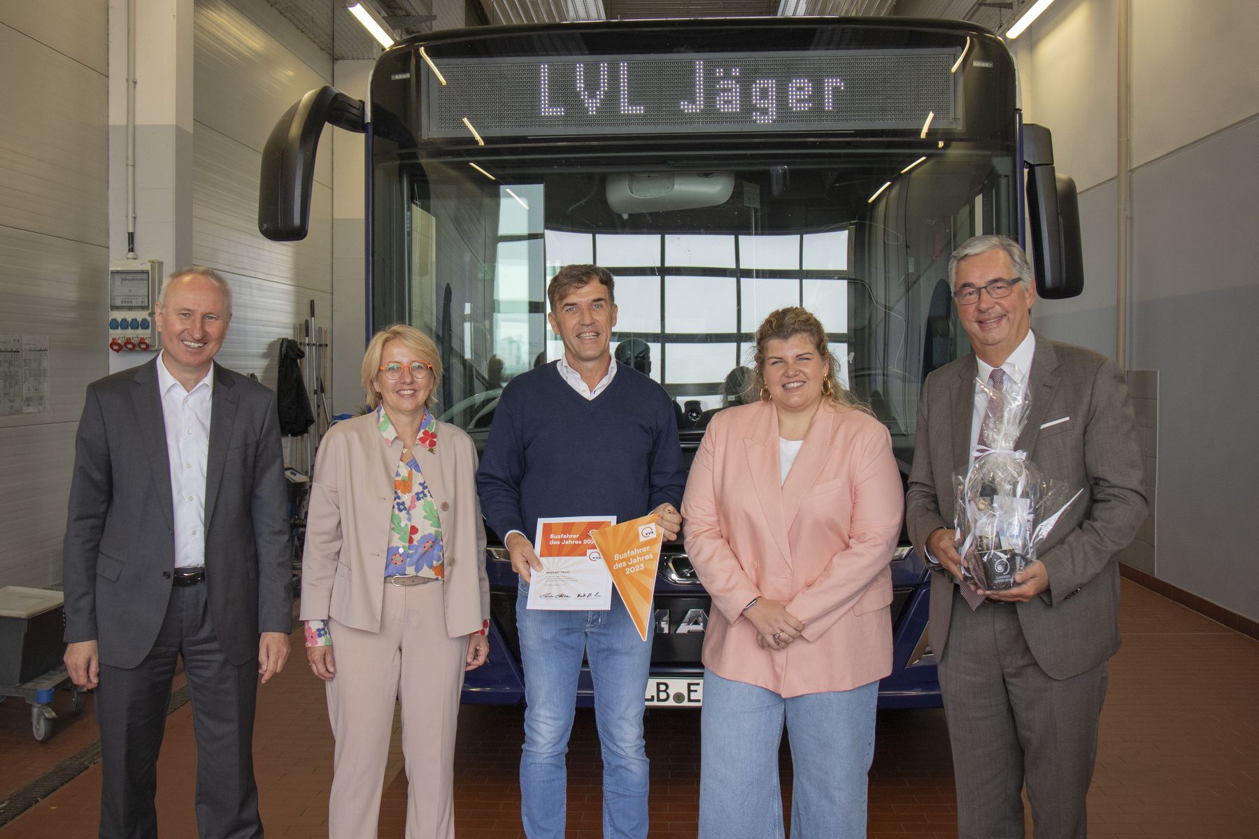 Fünf Personen stehen vor einem Bus mit der Aufschrift „LVL Jäger“. Sie lächeln und halten Urkunden oder Preise in den Händen. Die Szene zeigt eine feierliche Übergabe oder Auszeichnung im Transportbereich.