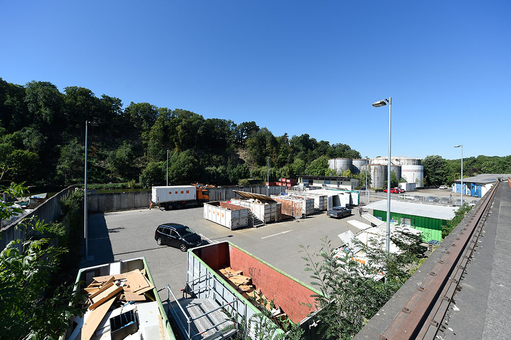 Ein großer Parkplatz mit mehreren Containern für Abfälle, umgeben von Bäumen. Im Hintergrund sind einige Lagerbehälter sichtbar. Der Himmel ist klar und blau, es ist Tageslicht. Ein Auto steht auf dem Platz, während die Umgebung ruhig wirkt.