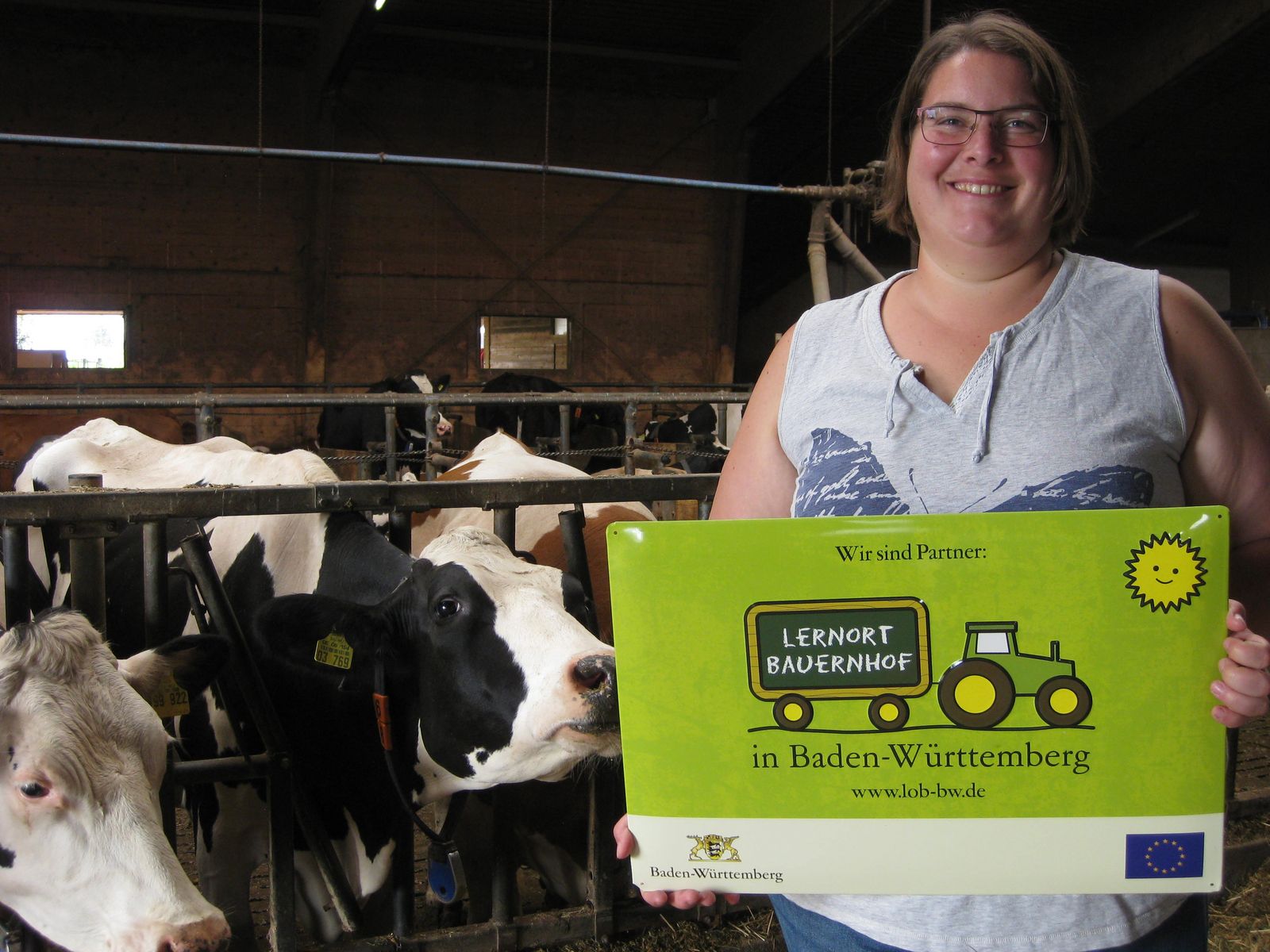 Una donna sorridente tiene un cartello verde che recita "Lernort Bauernhof in Baden-Württemberg" accanto a mucche in un fienile. La scena rappresenta un'iniziativa educativa legata all'agricoltura.