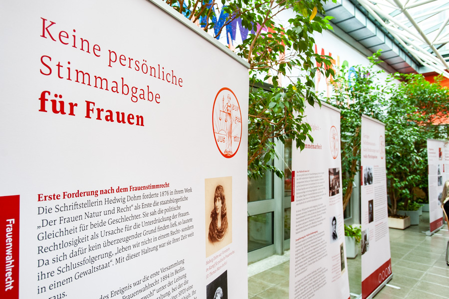 Immagine di una mostra con pannelli informativi che trattano il tema dei diritti di voto per le donne. I pannelli includono testi in tedesco e mostrano fotografie storiche. Sullo sfondo si vedono piante in un ambiente interno.