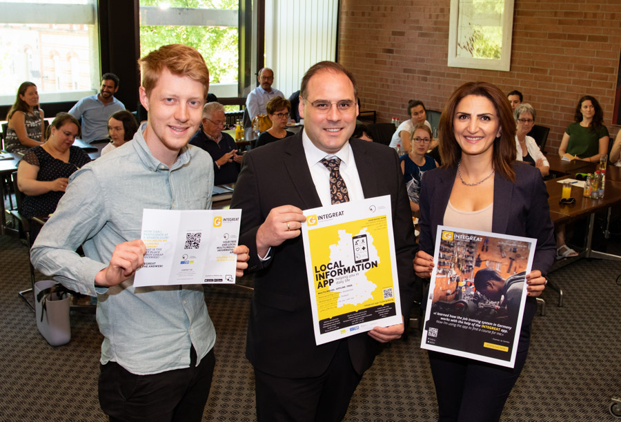 Three individuals stand together, each holding promotional materials for a local information app. They are in a crowded room with an audience in the background. The setting suggests a community event focusing on local engagement and resources.