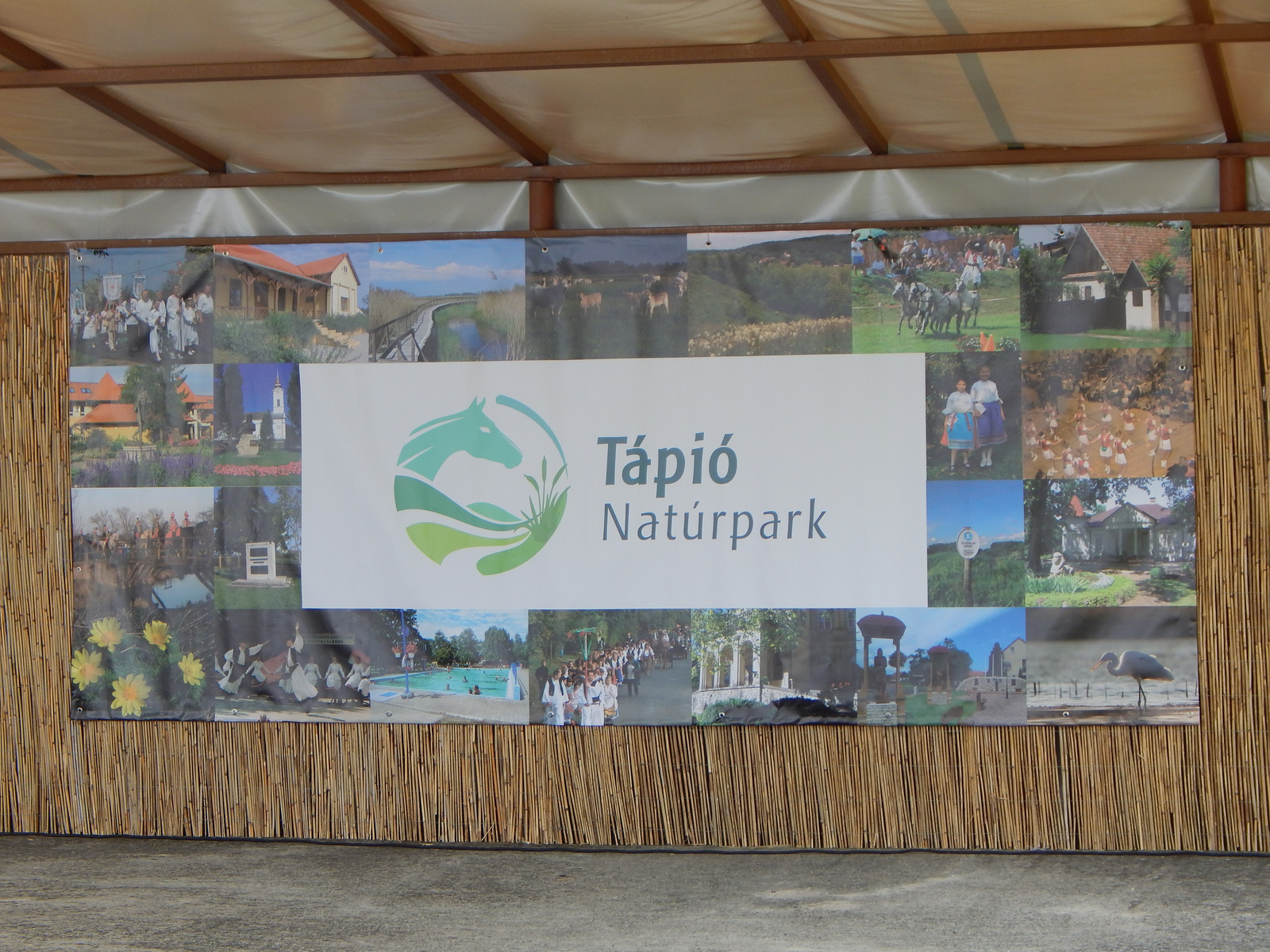 Ein großes Banner mit dem Text "Tápió Naturpark" in der Mitte. Umgeben von verschiedenen Bildern, die Landschaften, Tiere und Aktivitäten im Naturpark zeigen. Das Banner ist an einer Holzverkleidung angebracht und vermittelt den Eindruck eines einladenden Naturerlebnisses.