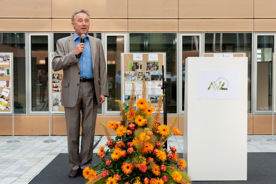 A man in a gray suit stands on a stage, holding a microphone and speaking to an audience. In the background, there are photo displays, and in front of him, a floral arrangement with orange flowers is visible. A podium with a logo is positioned beside him.