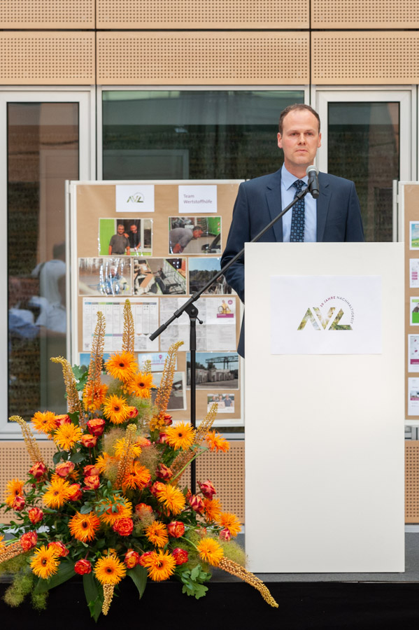 A man in a suit speaks at a podium during an event. He stands before a display board featuring various photographs and information. In front of him, a bouquet of vibrant orange and yellow flowers adds color to the setting.