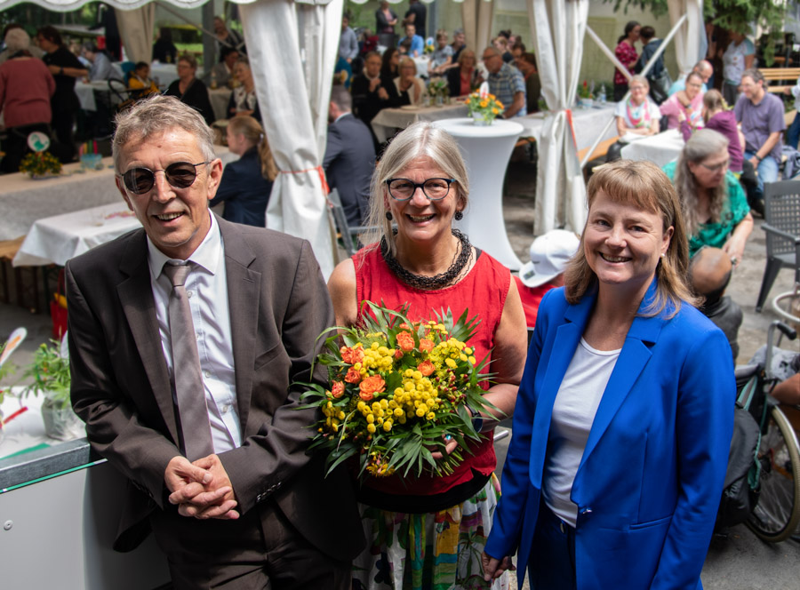 Drei Personen stehen im Vordergrund: ein Mann in dunklem Anzug und Sonnenbrille, eine Frau mit Brille und buntem Kleid, die einen Blumenstrauß hält, sowie eine Frau in blauem Blazer. Im Hintergrund sind viele Menschen bei einer Veranstaltung zu sehen.