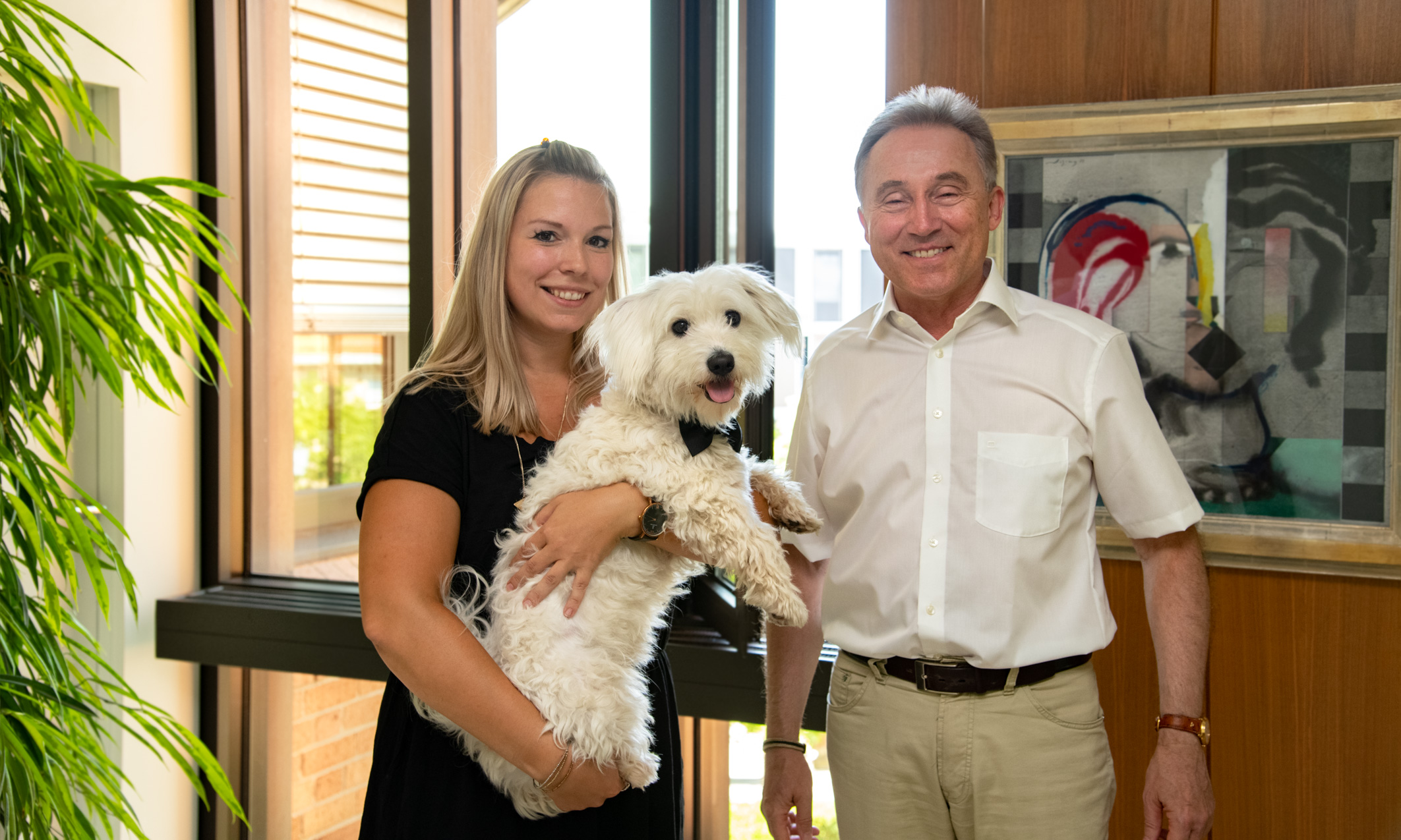 Eine Frau hält einen kleinen, weißen Hund in ihren Armen. Neben ihr steht ein Mann in heller Kleidung. Beide lächeln freundlich. Im Hintergrund sind große Fenster und ein modernes Kunstwerk zu sehen.
