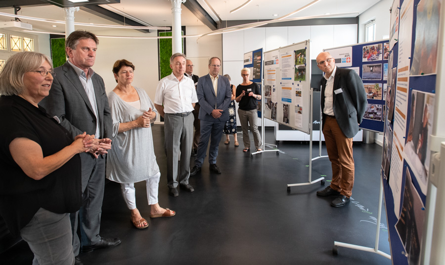 Eine Gruppe von fünf Personen steht in einem modernen Raum vor Informationsplakaten, die an Ständern ausgestellt sind. Einige Teilnehmer unterhalten sich, während sie auf die Plakate zeigen. Der Raum wirkt hell und einladend.
