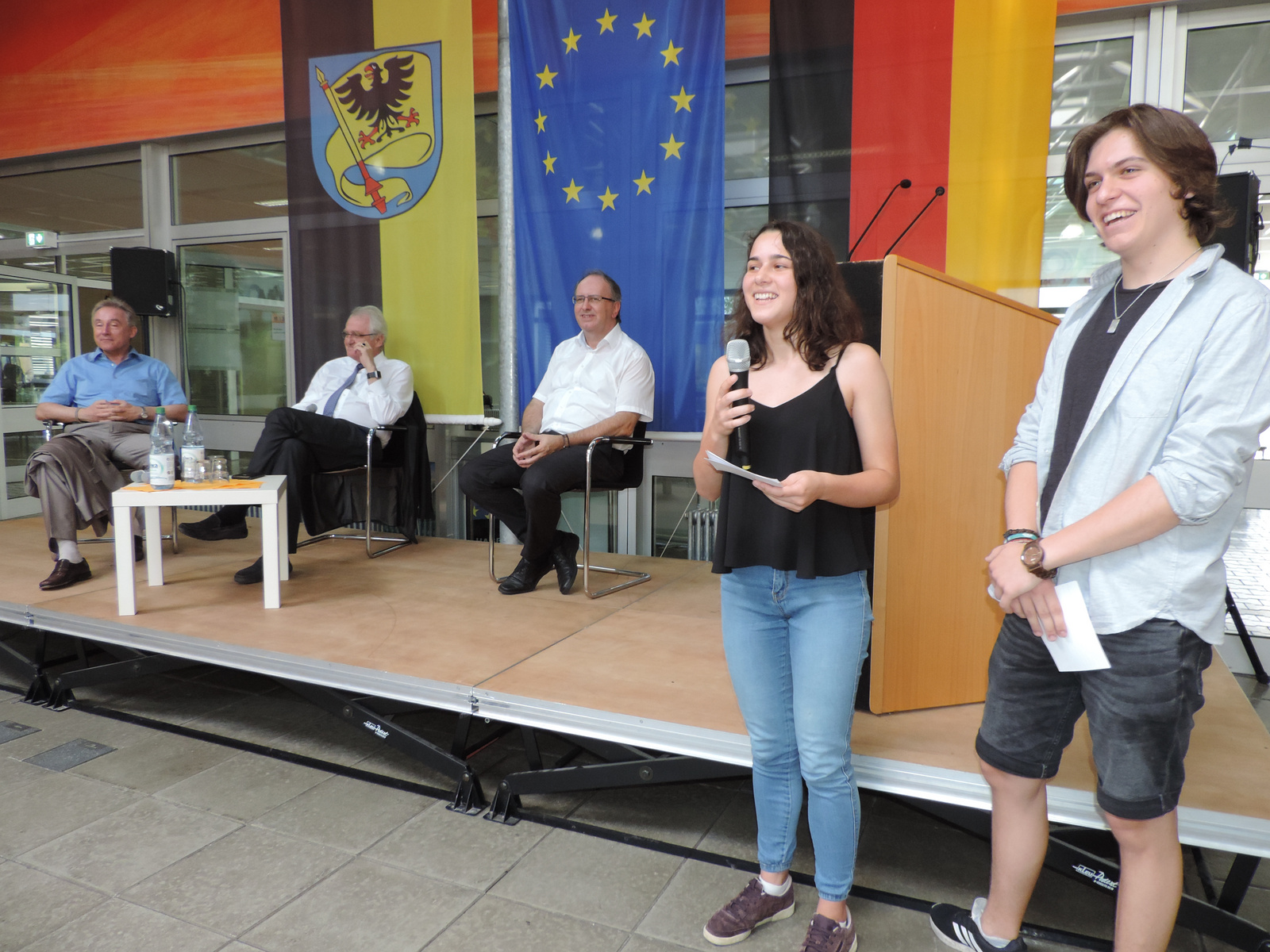 A young woman stands on stage speaking into a microphone, while three seated older men listen. The backdrop features flags of Germany and the European Union, suggesting the event focuses on civic engagement or political discussion. A wooden podium is present on stage.