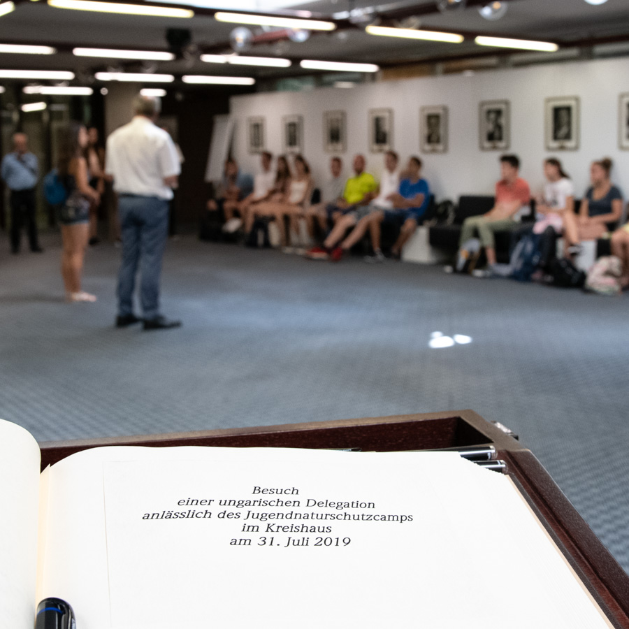 Eine Gruppe von Menschen sitzt und steht in einem Raum, während eine Präsentation stattfindet. Im Vordergrund liegt ein offenes Buch mit dem Titel „Besuch einer ungarischen Delegation anlässlich des Jugendnaturschutzcamps am 31. Juli 2019“.