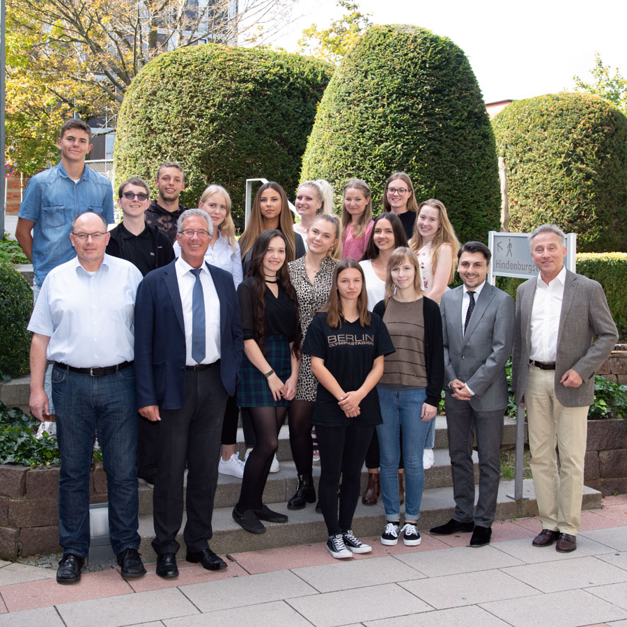 Eine Gruppe von 20 Menschen steht im Freien vor einer grünen Hecke. Die Gruppe besteht aus verschiedenen Altersgruppen und Geschlechtern, einige in formeller Kleidung und andere leger. Es ist ein sonniger Tag, und die Personen lächeln für das Foto.