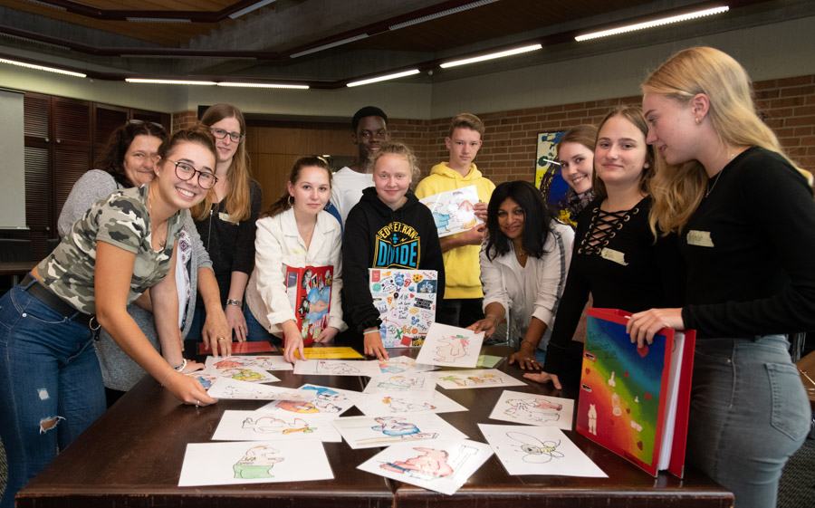 Eine Gruppe von Schülern steht um einen Tisch, auf dem zahlreiche bunte Zeichnungen liegen. Einige Schüler halten ihre eigenen Kunstwerke in den Händen. Die Atmosphäre ist freundlich und die Teilnehmer scheinen stolz auf ihre kreativen Arbeiten zu sein.